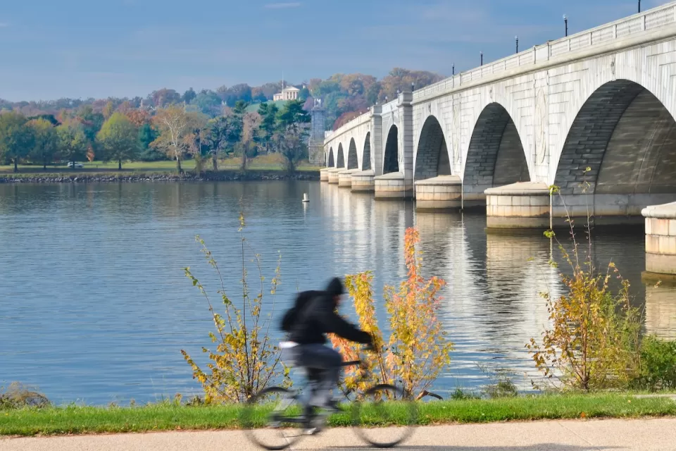 Person on bike on path next to Memorial Bridge in Washington, D.C. with fall foliage in background across river.